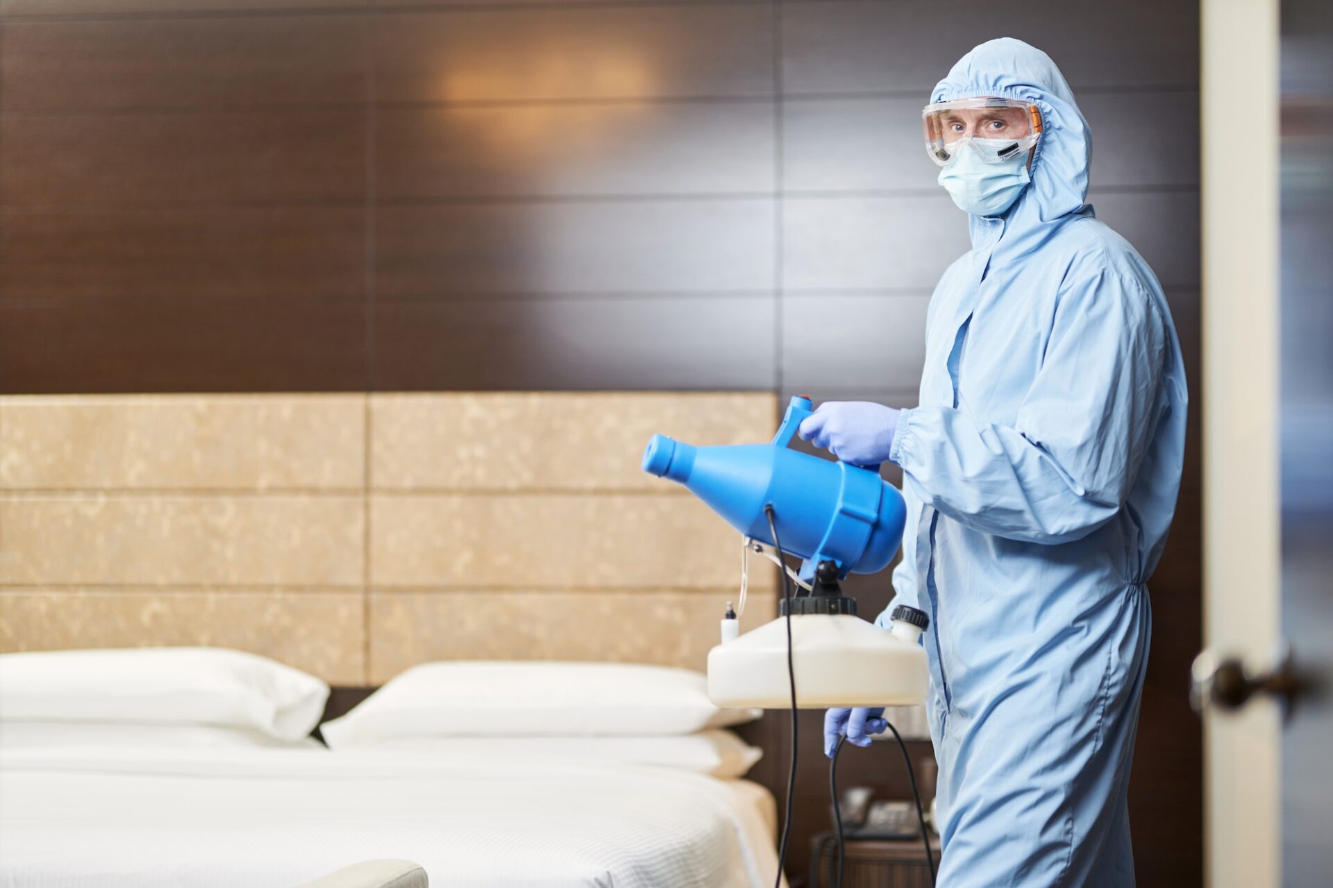 Man in protective clothes standing with disinfectant near bed in hotel room